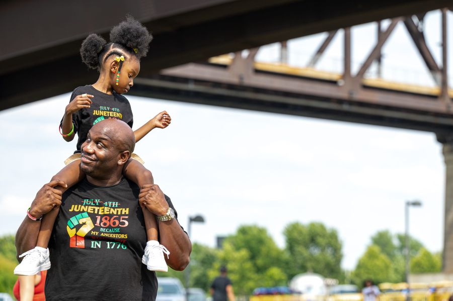 Dad holds little girl on his shoulders. They wear matching Juneteenth tees
